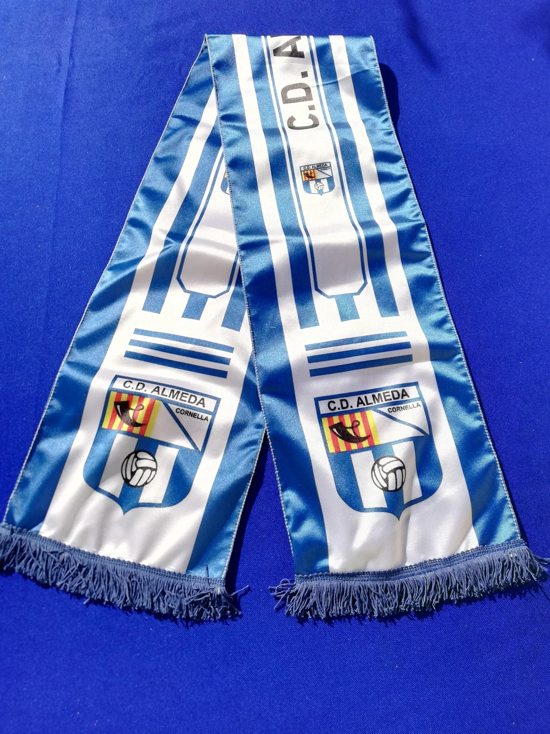 Bufanda deportiva a rayas azules y blancas con extremos con flecos y escudos al estilo argentino sobre fondo azul.