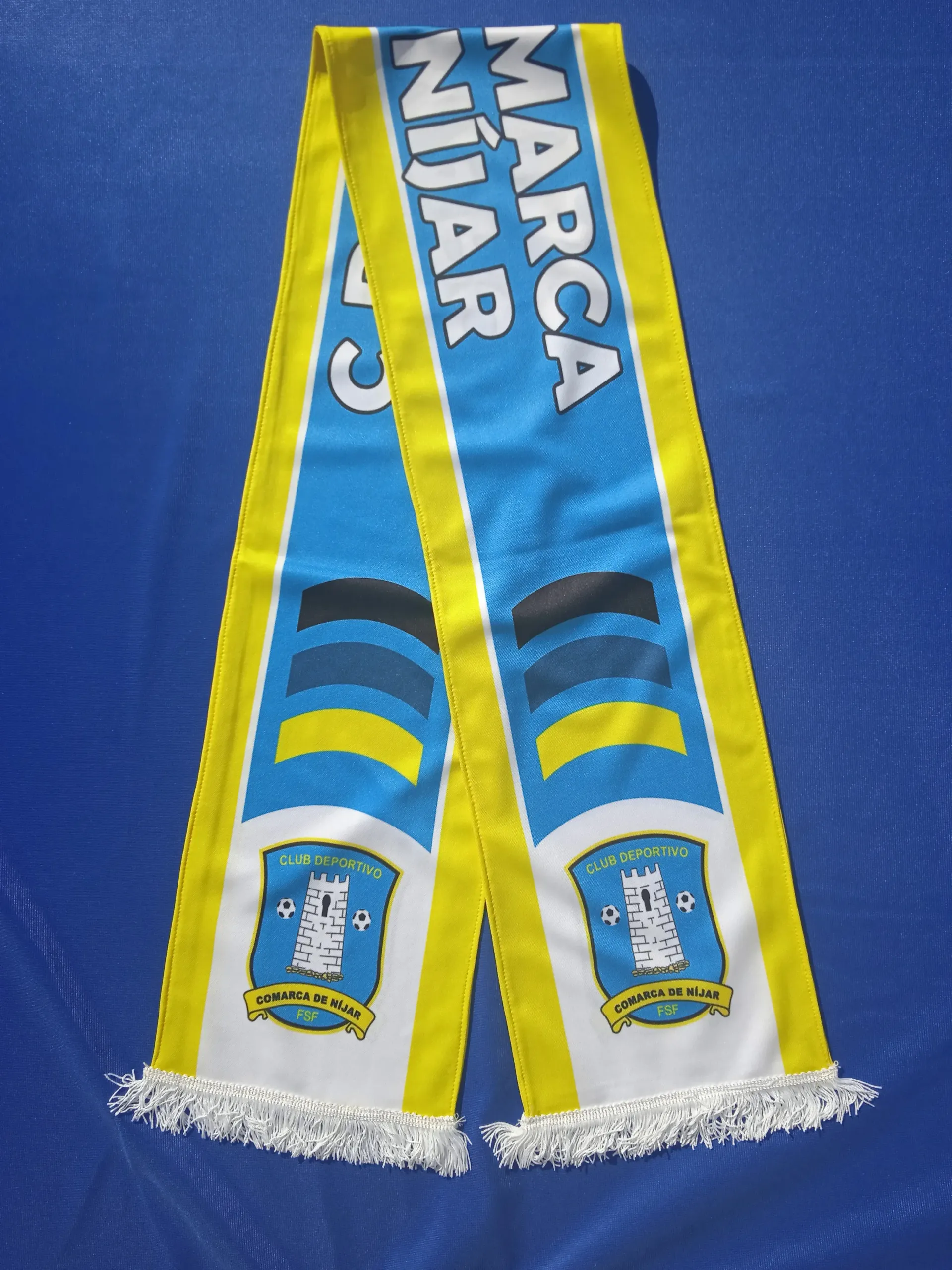 Bufanda Manta Sports azul y amarilla con flecos blancos, extendida sobre un fondo azul.