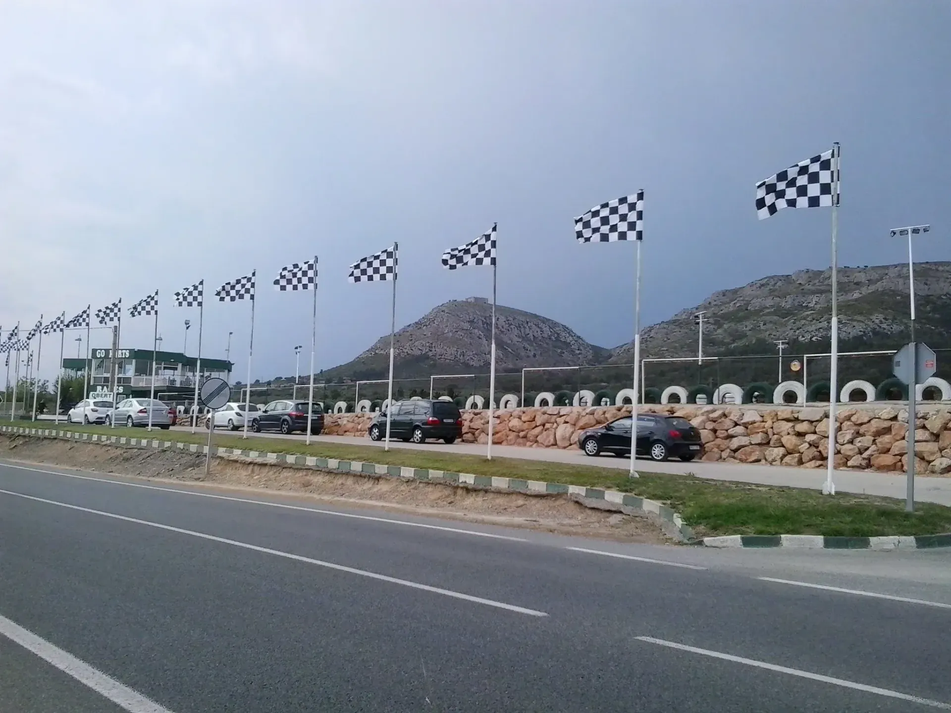 Banderas a cuadros bordean una carretera cerca de coches aparcados y colinas bajo un cielo nublado.