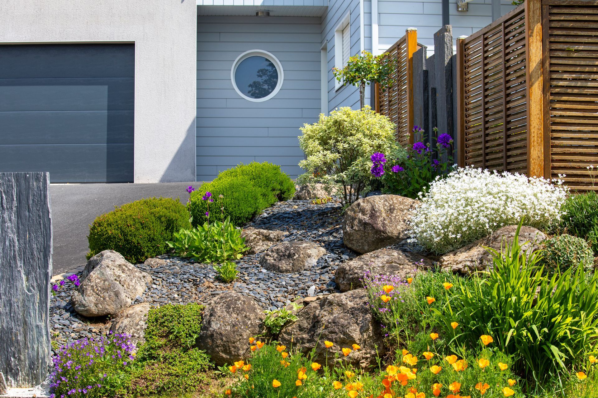 Cour avant paysagée avec rochers, verdure et fleurs à côté d'un bâtiment moderne avec une porte de garage.