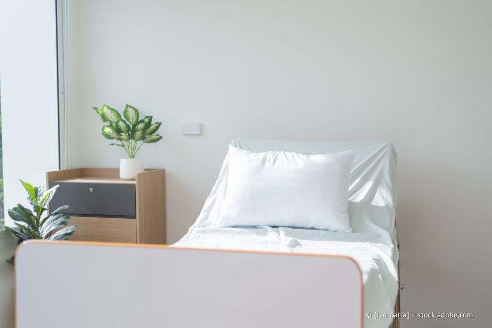 Krankenhausbett mit weißer Bettwäsche, Kissen und Nachttisch. Grüne Pflanze am Fenster.