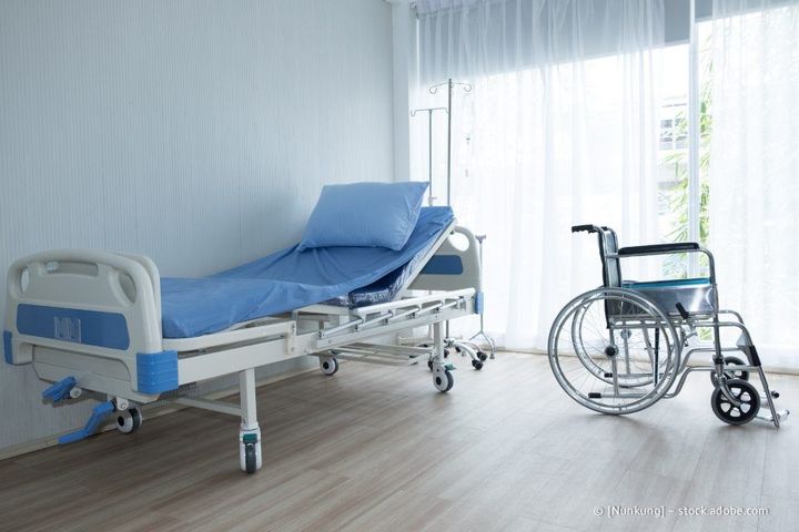 Krankenzimmer mit Bett und Rollstuhl neben einem großen Fenster.