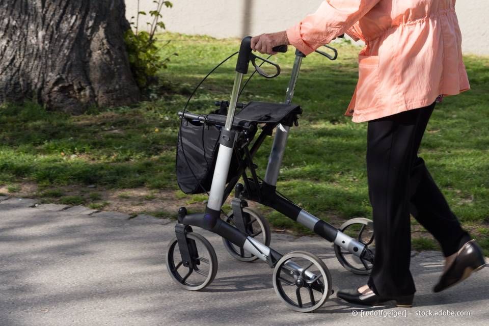 Ältere Person, die mit einem Rollator auf einem Bürgersteig geht; sie trägt schwarze Hosen und eine lachsfarbene Jacke.