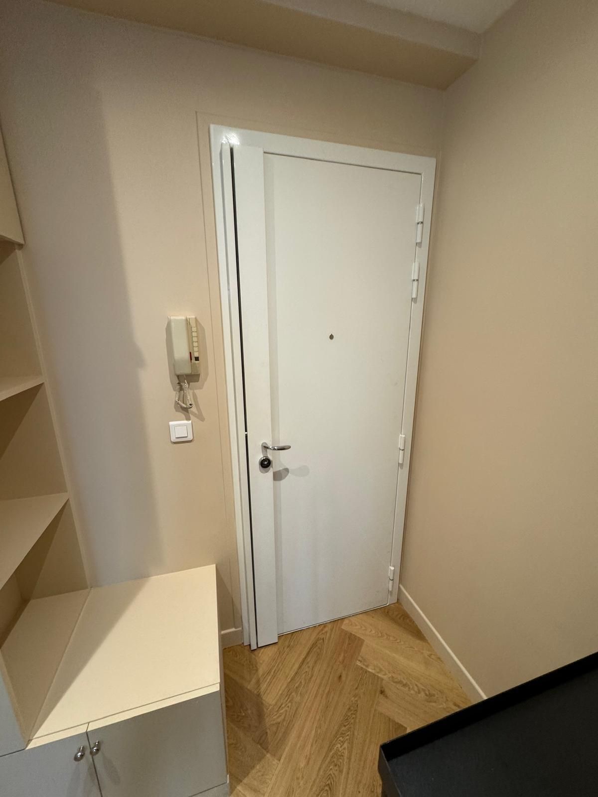Petite entrée beige avec une porte blanche, un interphone mural et un parquet à chevrons.