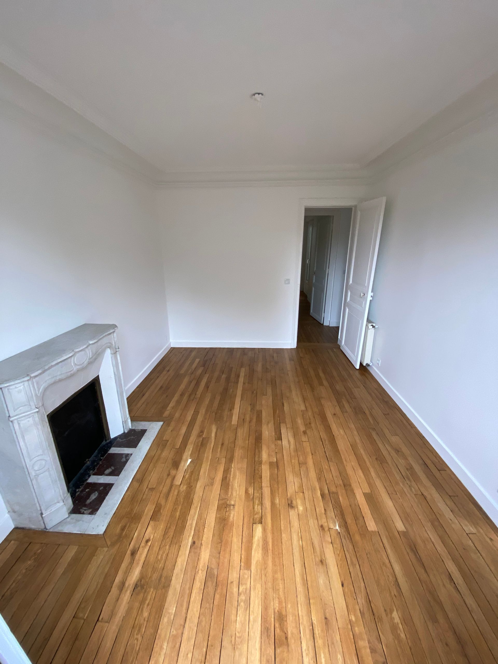 Salon vide et blanc, avec un parquet et une cheminée sur la gauche.