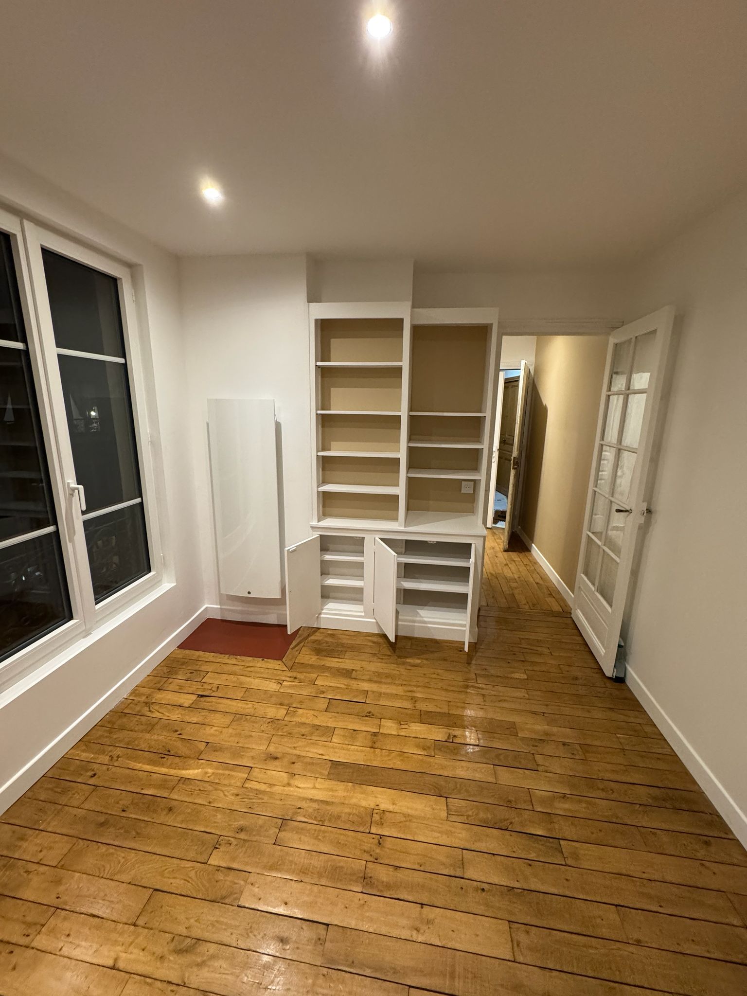 Vue intérieure d'une pièce avec parquet ciré, murs blancs, étagères intégrées, radiateur et porte vitrée.