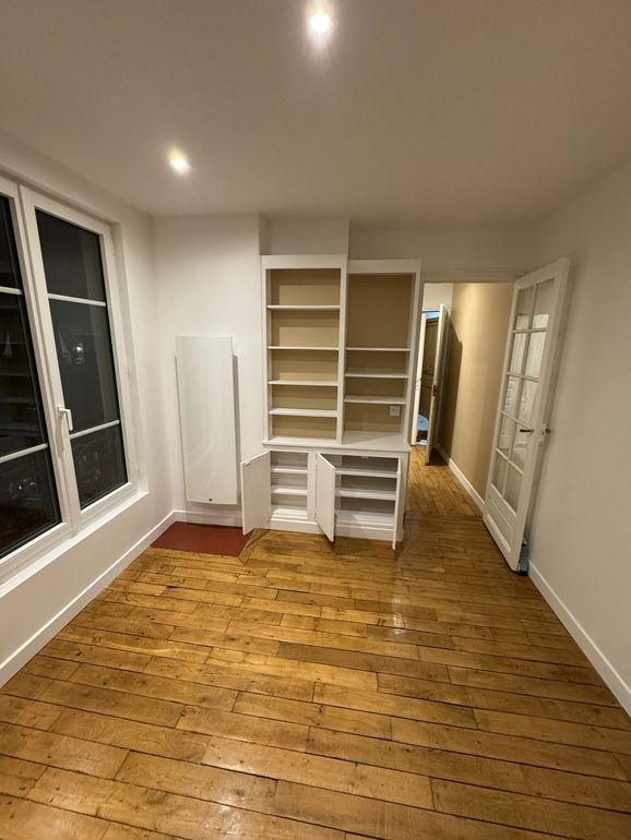 Vue intérieure d'une pièce avec parquet ciré, murs blancs, étagères intégrées, radiateur et porte vitrée.