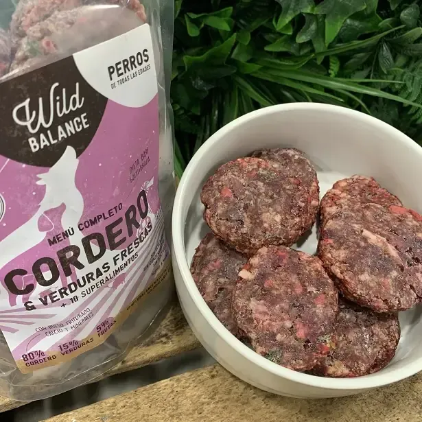 Paquete y cuenco de comida cruda de cordero para perros, junto a vegetación.