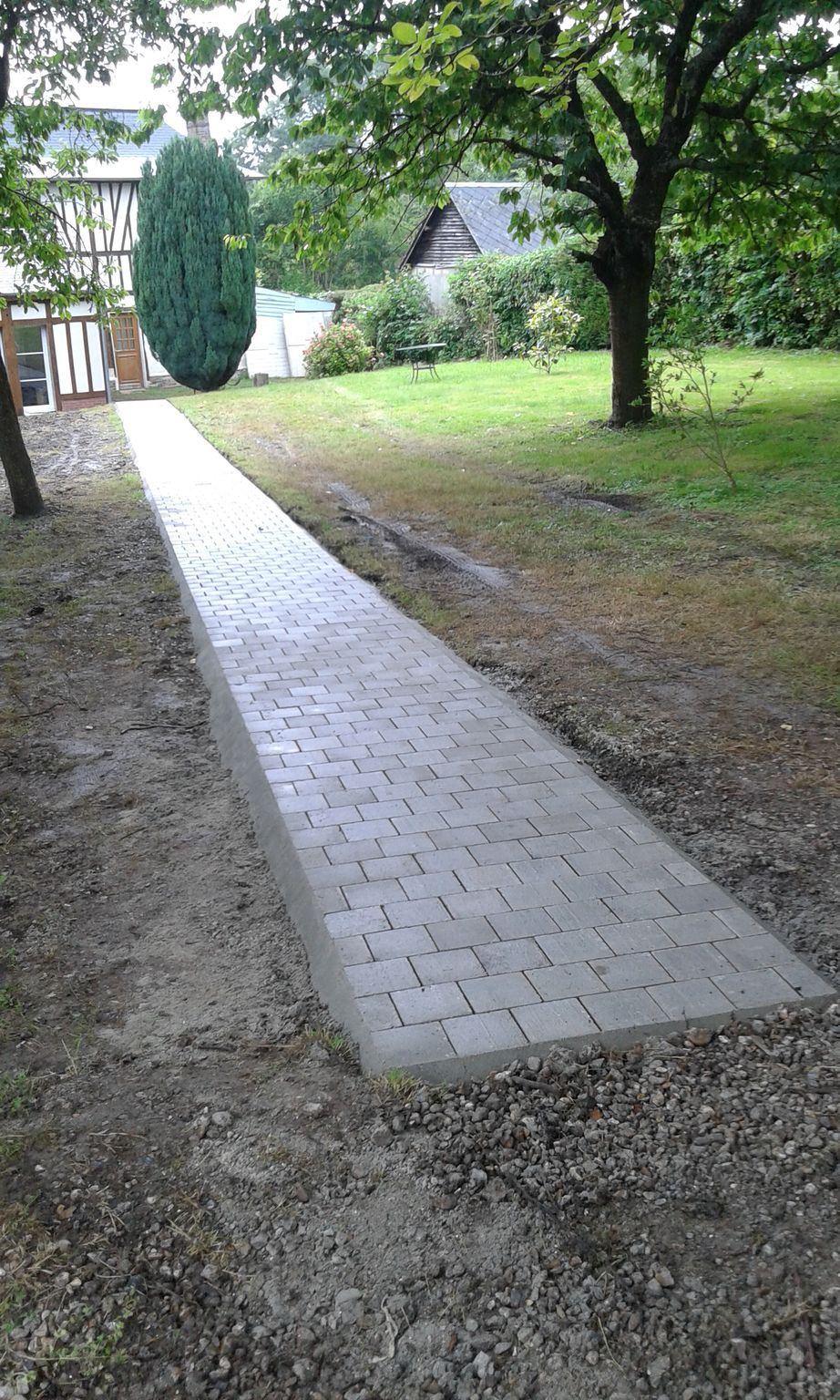 Allée de briques traversant une cour herbeuse, menant vers un portail et des arbres.