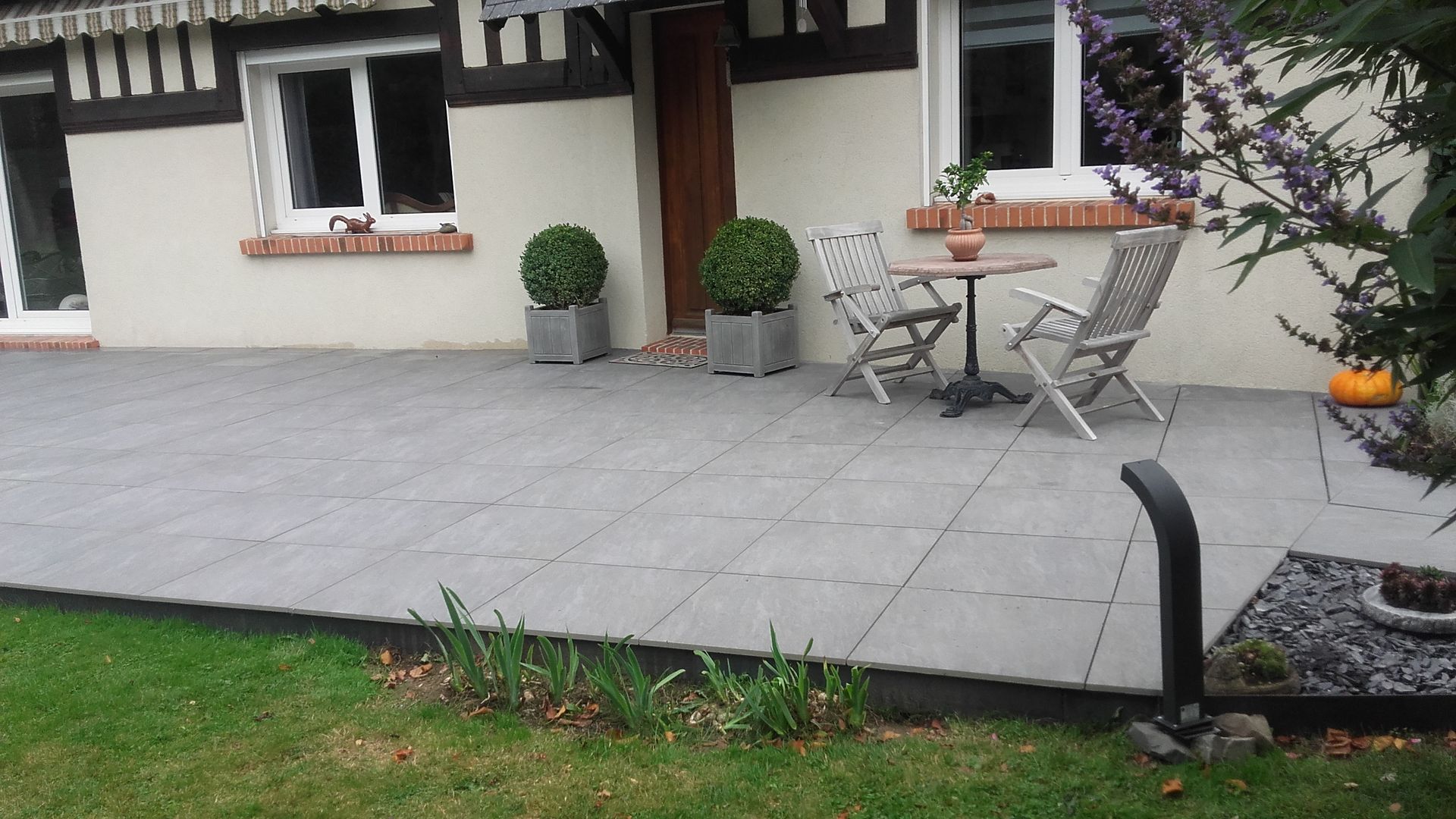 Terrasse carrelée grise avec mobilier de jardin, plantes en pot et pelouse.