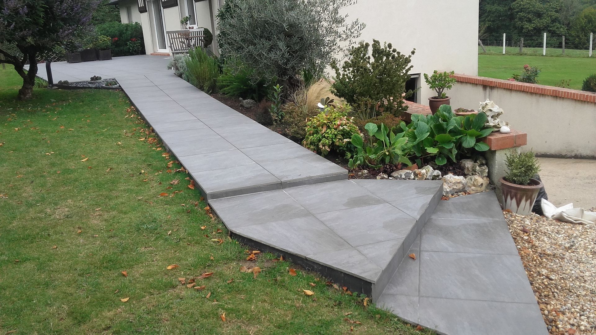 Un chemin carrelé de gris et des marches mènent à un bâtiment, bordé de verdure de part et d'autre.