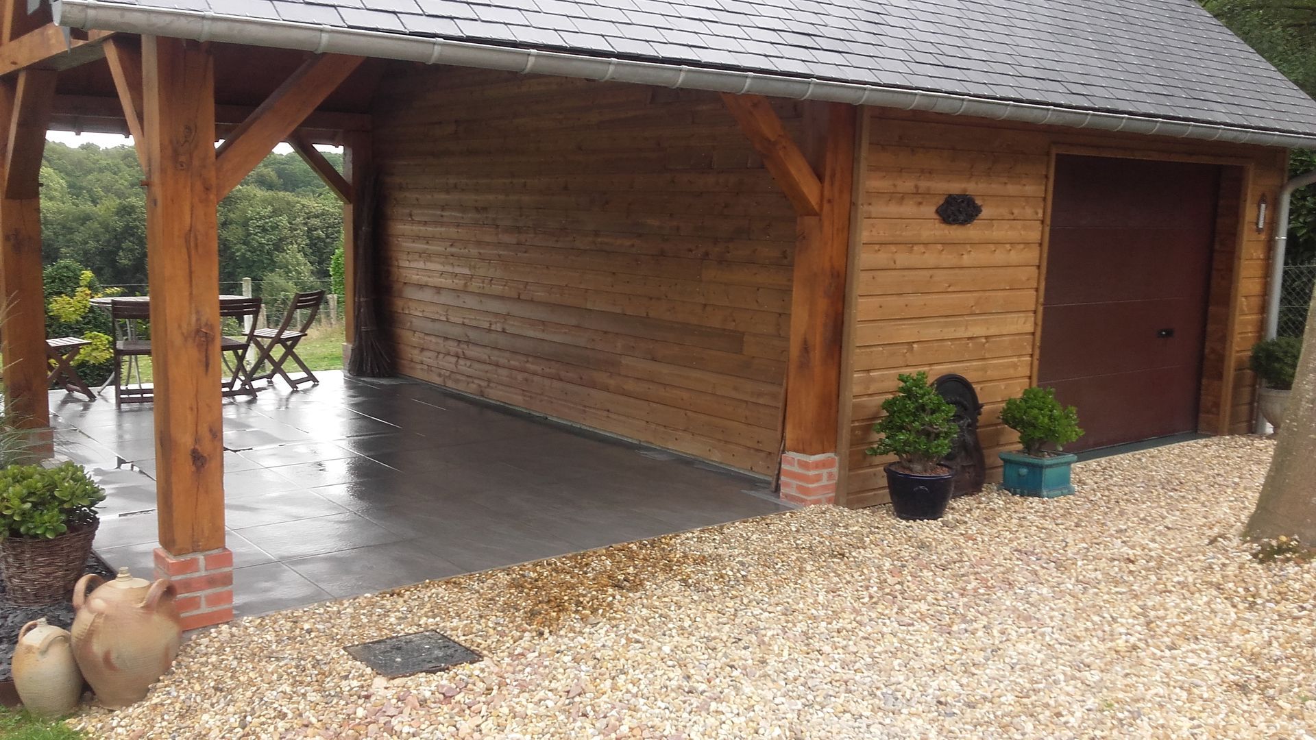 Abri de jardin en bois avec porche, allée de gravier et plantes. Porte et bardage marron, toit gris.