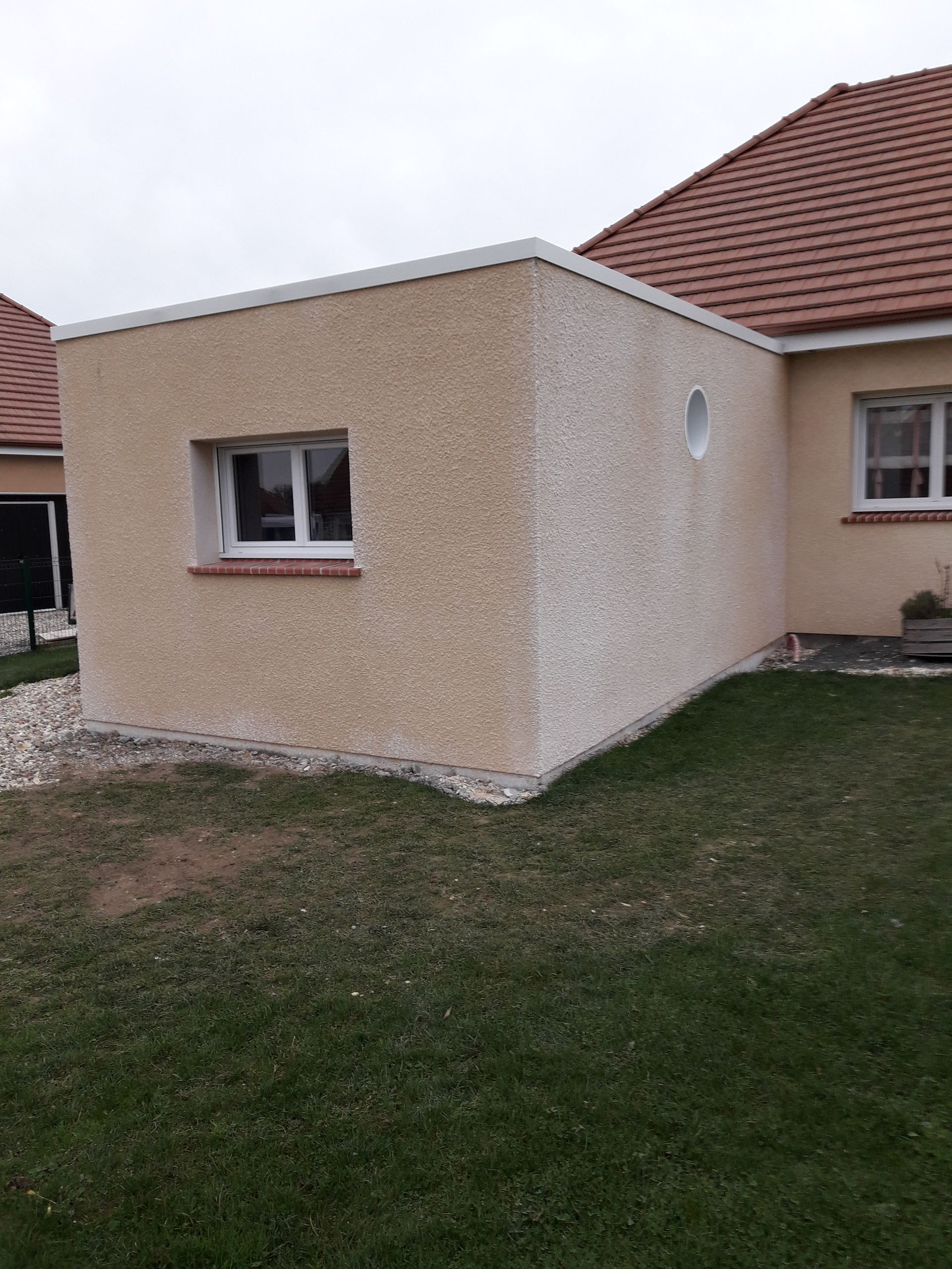 Extension extérieure beige avec une fenêtre, attenante à une maison au toit de tuiles orange, sur une pelouse.