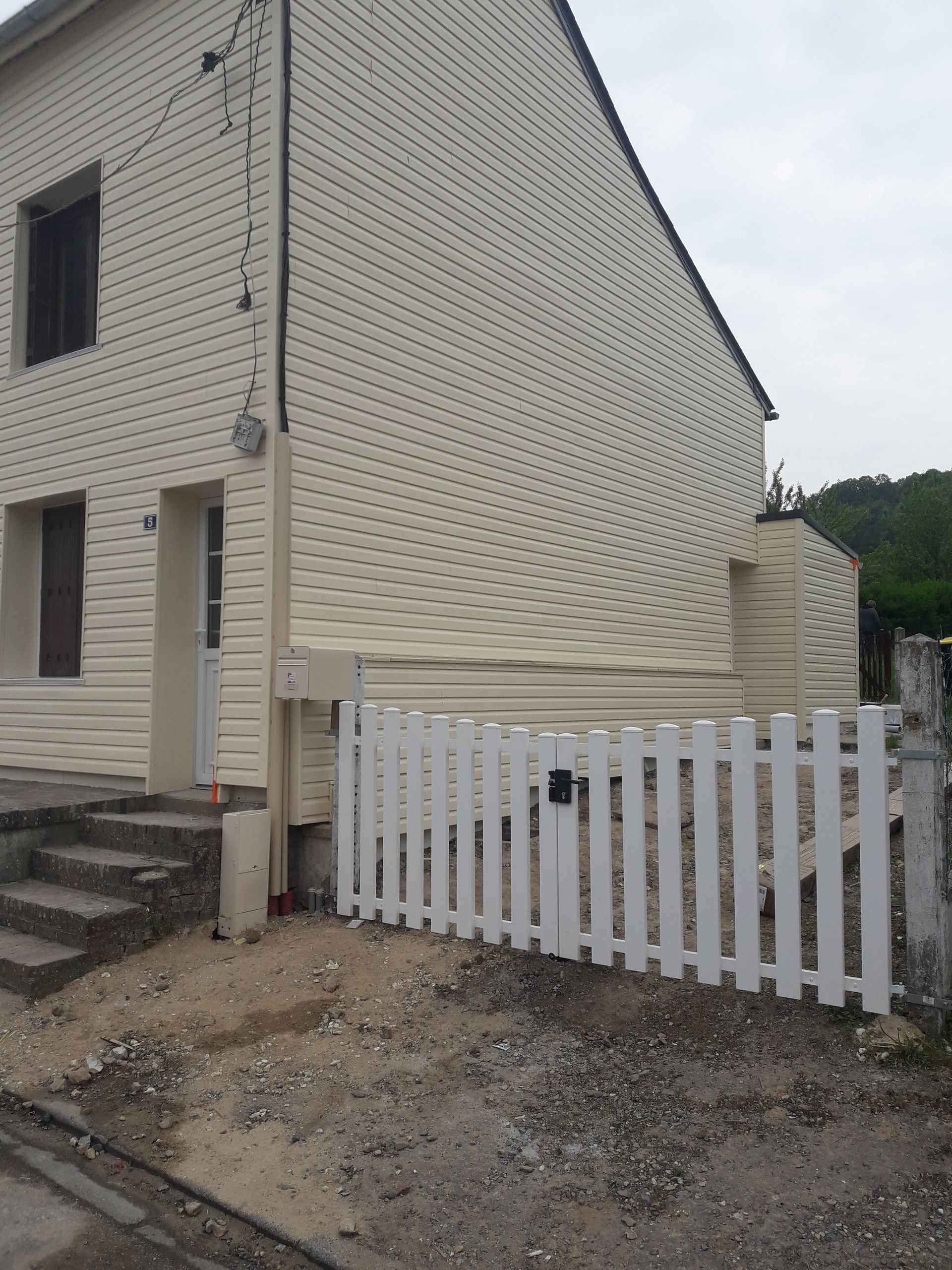 Maison beige avec une clôture blanche et des marches menant à la porte d'entrée, sur un chemin de terre.
