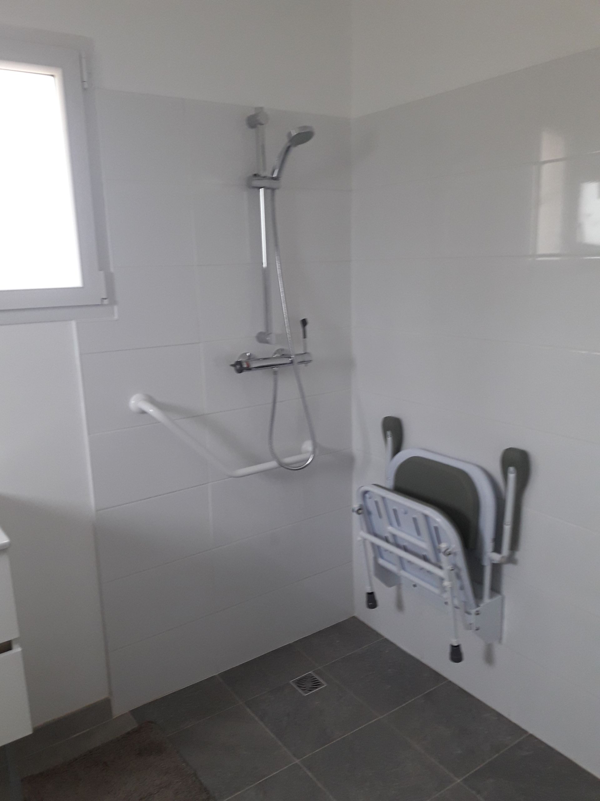 Douche accessible avec siège rabattable, barres d'appui et pommeau de douche réglable dans un angle. Murs blancs, sol gris.