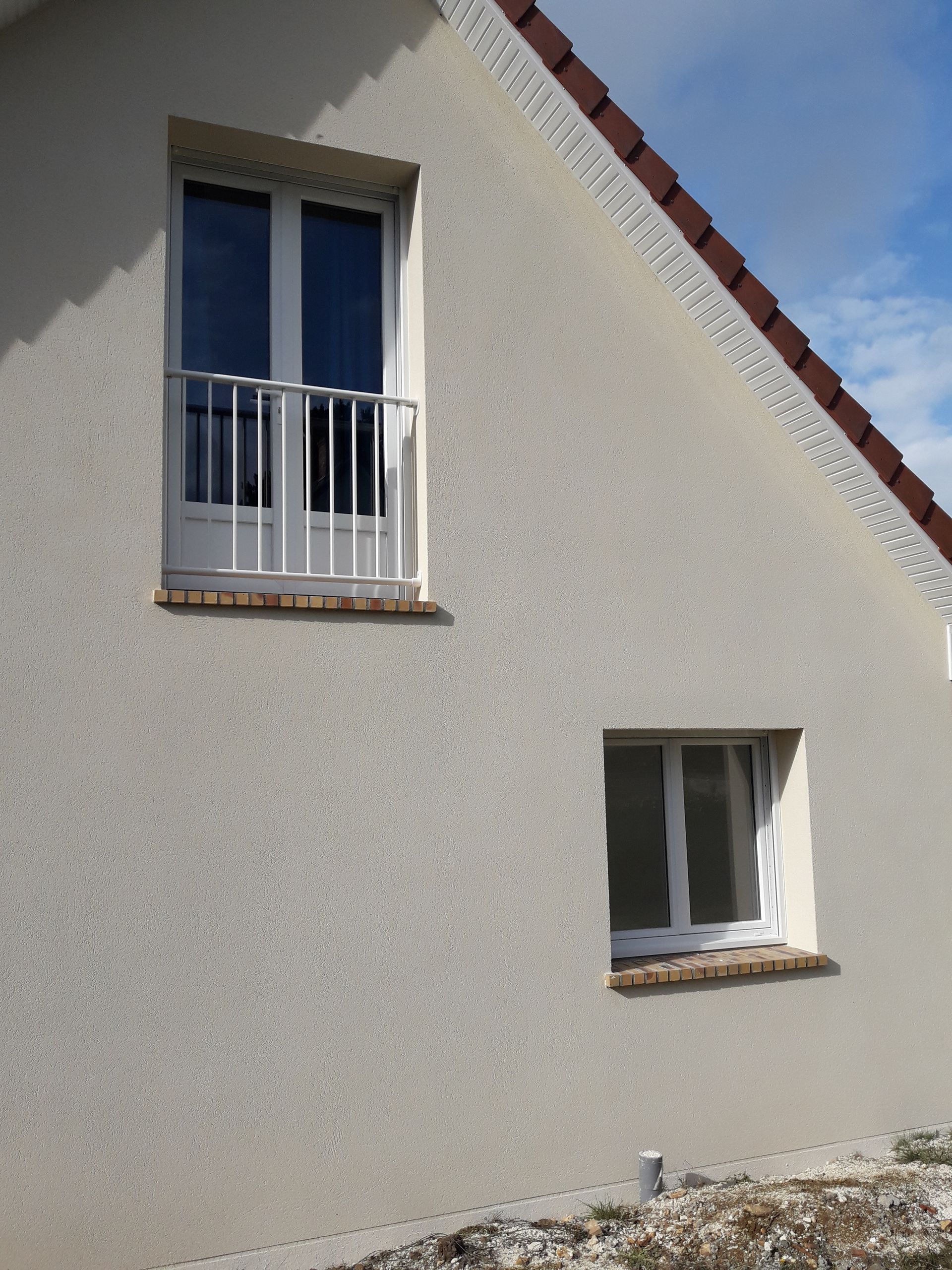Extérieur d'une maison avec deux fenêtres à cadre blanc, dont une avec un petit balcon. Revêtement extérieur en stuc gris, toit de tuiles rouges.