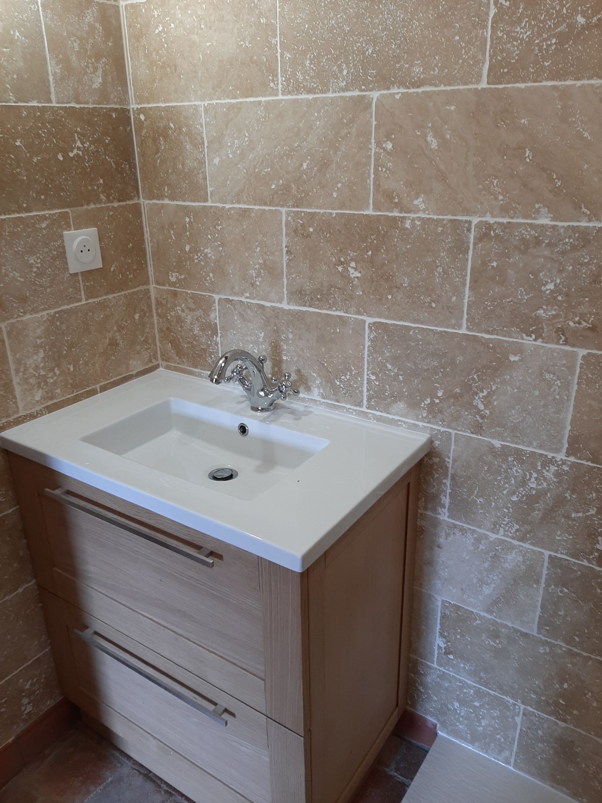 Salle de bains avec murs carrelés de carrelage en pierre beige, meuble-lavabo en bois clair avec comptoir blanc et robinet chromé.