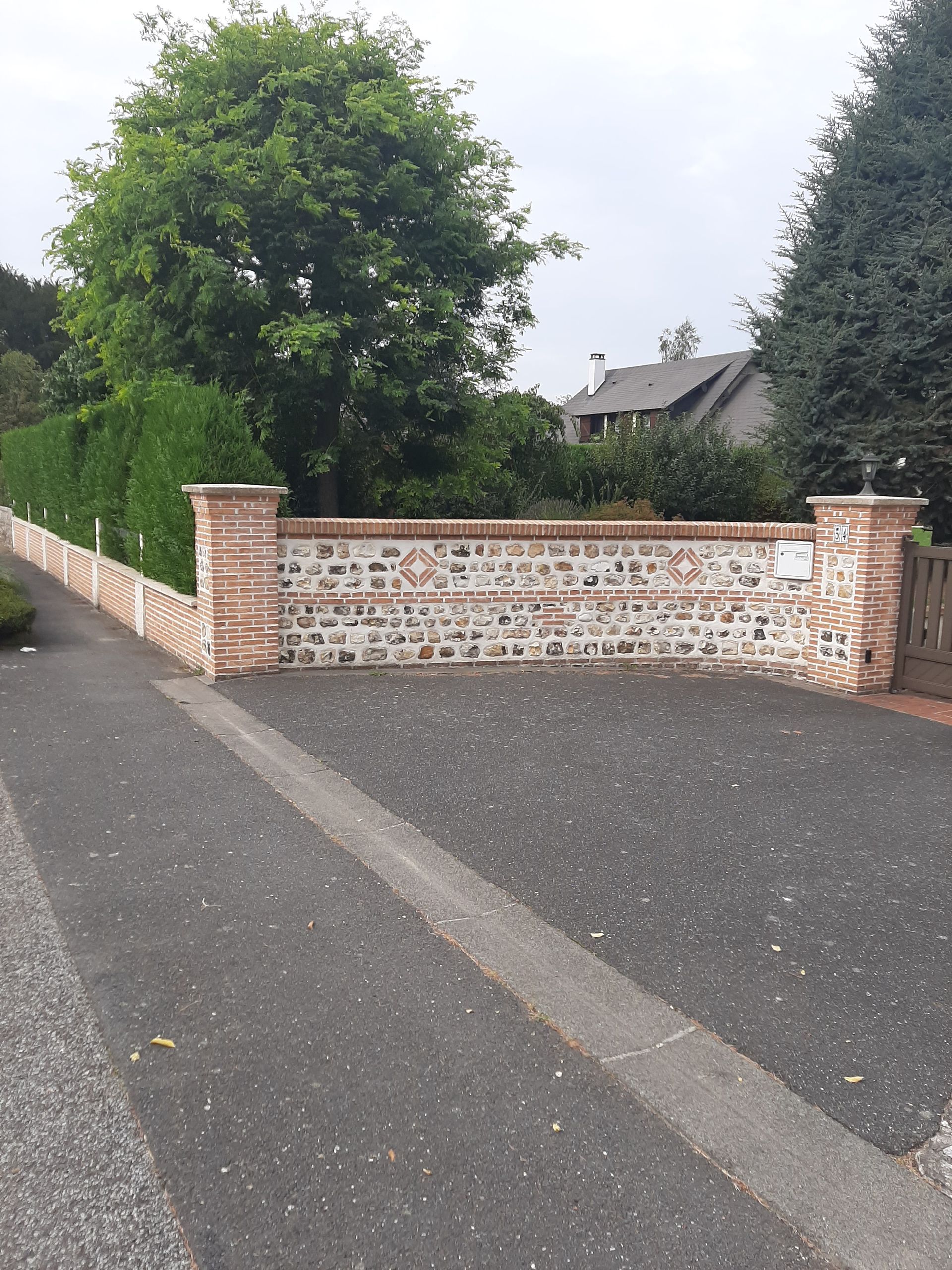 Mur en briques et en pierres avec haie, arbres et route asphaltée.
