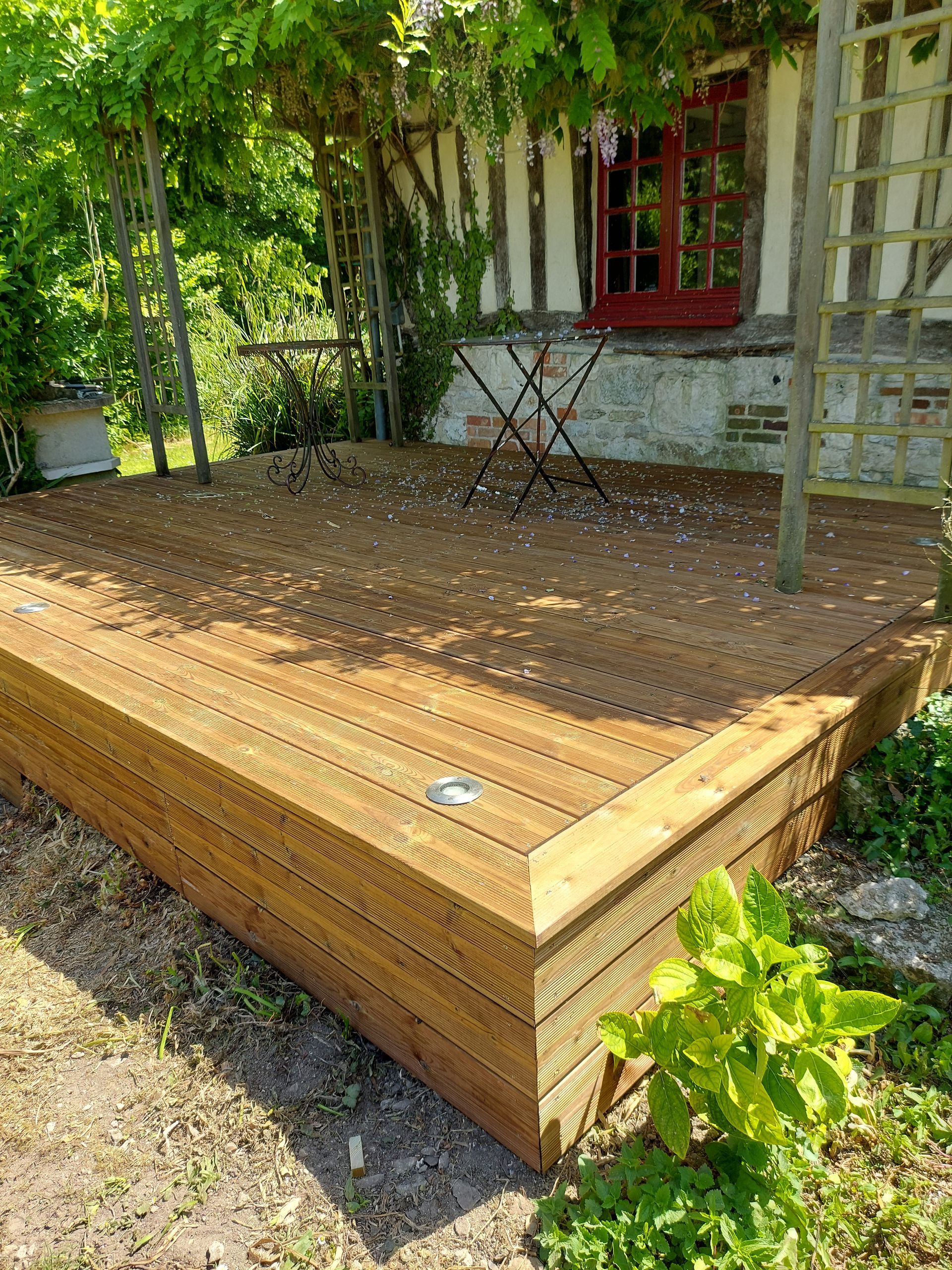 Terrasse en bois dans un jardin, avec une table et une maison en arrière-plan avec une fenêtre rouge.