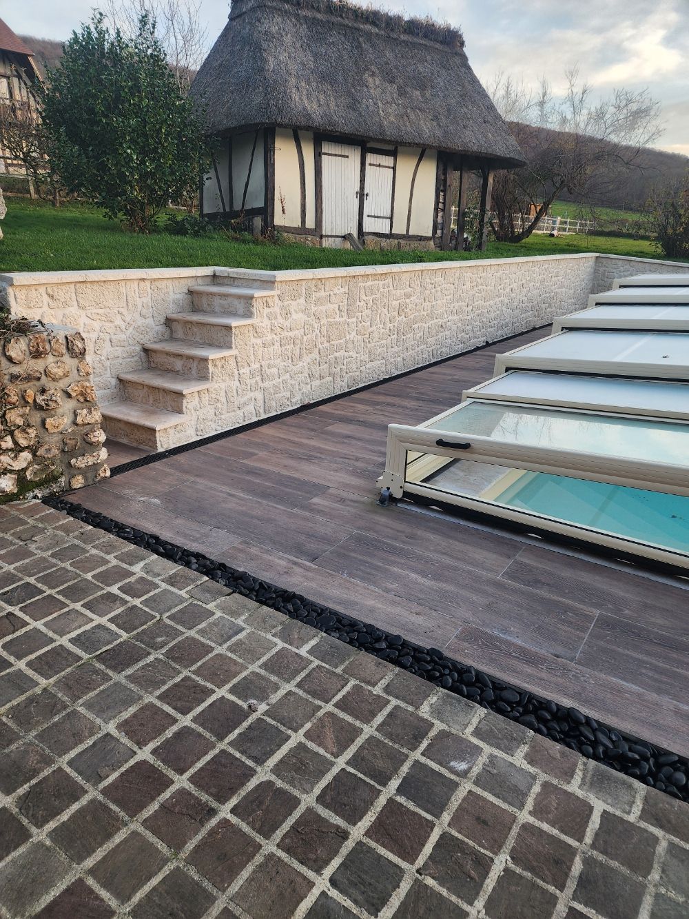 Terrasse de piscine avec mur et marches en pierre, terrasse en bois et bâtiment au toit de chaume en arrière-plan.