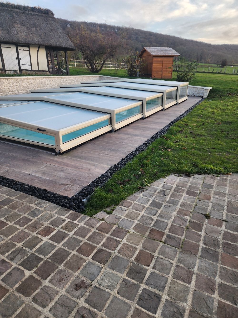 Piscine avec couverture rétractable dans un jardin, à côté d'une pelouse et d'une terrasse en pierre.