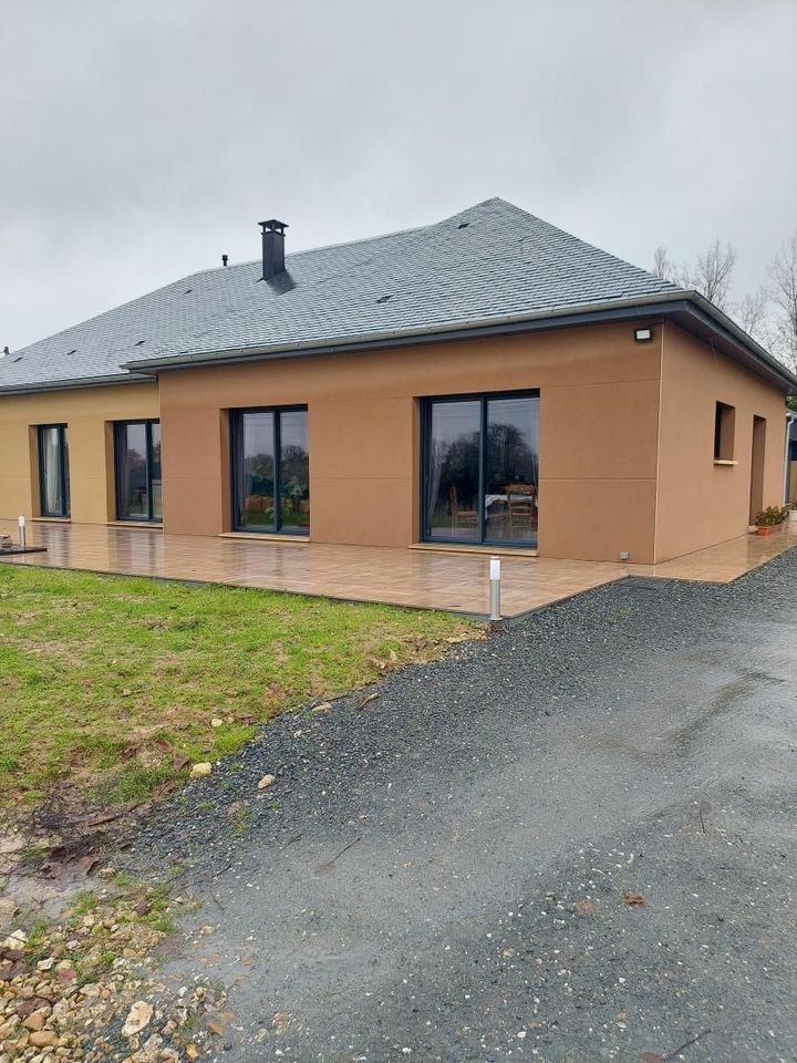 Maison moderne en stuc brun, avec des fenêtres à cadre foncé et un toit gris, par temps nuageux.