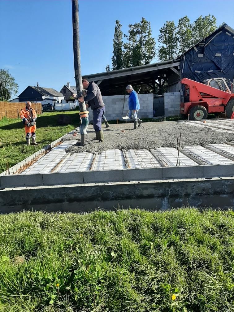 Des ouvriers coulent les fondations en béton d'un bâtiment ; tuyau de pompe, herbe verte, ciel bleu.