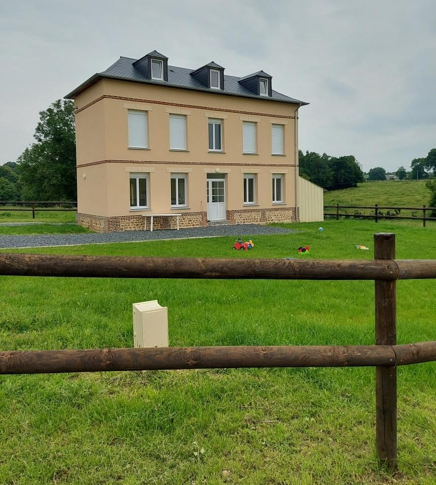 Maison beige à deux étages avec toit foncé et lucarnes, jouxtant un champ et une clôture en bois.