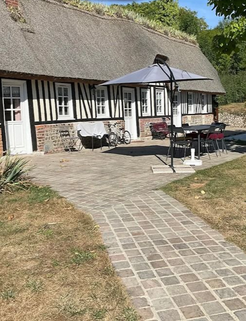 Une terrasse en briques mène à un chalet au toit de chaume avec un parasol, une table et des chaises.