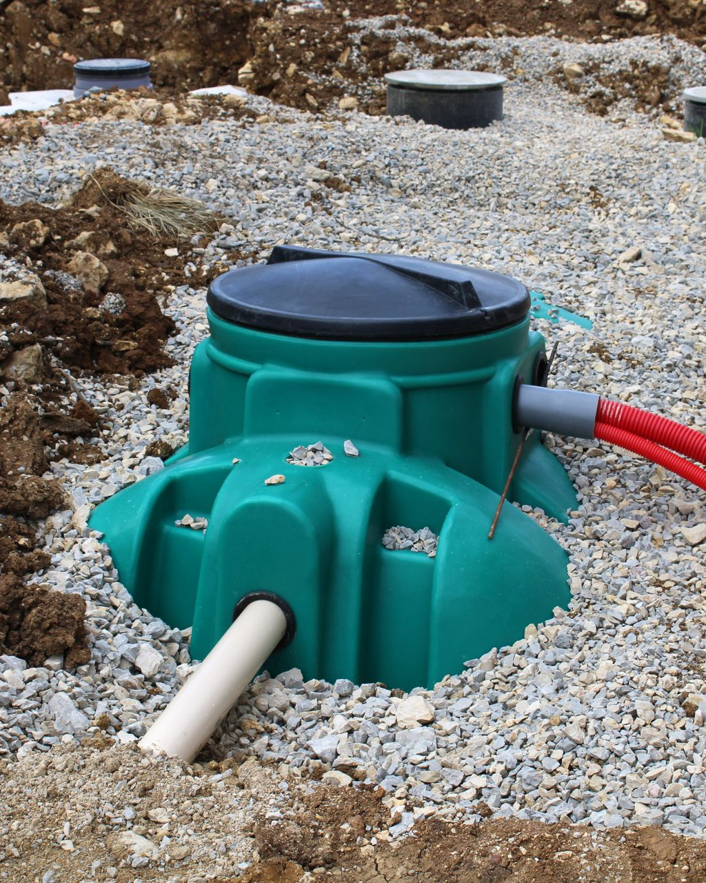 Fosse septique verte partiellement enterrée dans du gravier, avec couvercle noir et tuyaux en PVC.