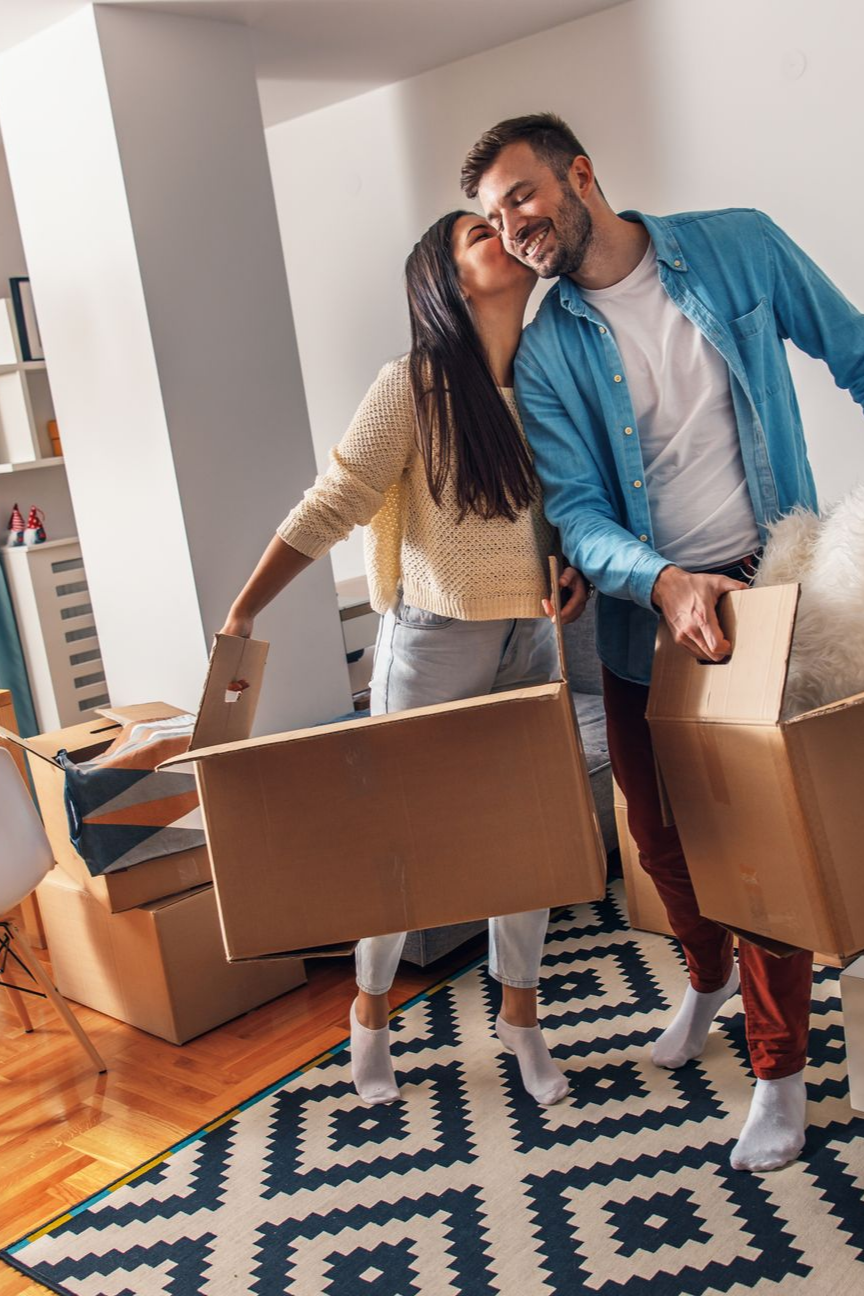 Un couple emménage, portant des cartons. La femme embrasse l'homme sur la joue en souriant. 