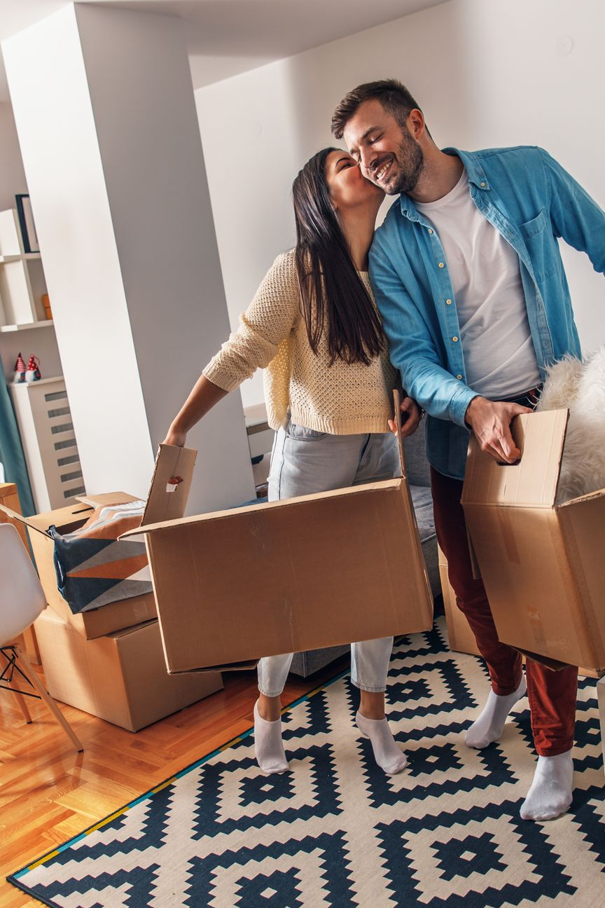 Un couple emménage dans sa nouvelle maison, portant des cartons. La femme embrasse l'homme sur la joue.