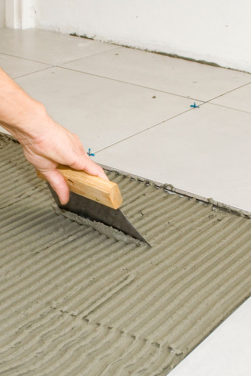 Utilisation manuelle d'une truelle crantée pour étaler du mortier gris sur un sol en vue de la pose de carrelage.