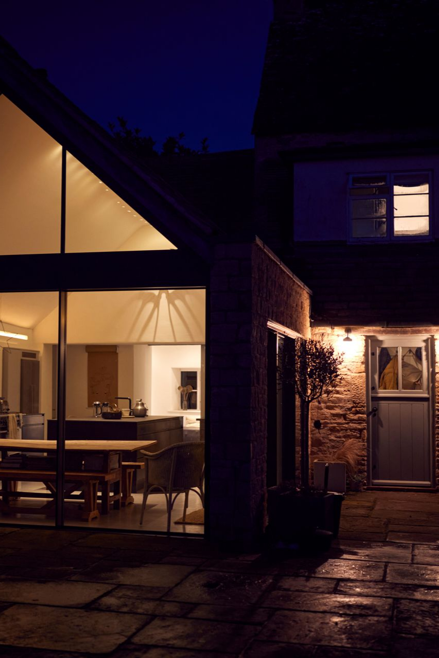 Vue nocturne d'une maison moderne aux grandes baies vitrées illuminant l'intérieur. Façade en briques, ciel bleu foncé.