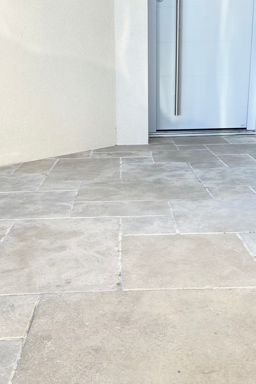 Le sol est carrelé de pierre et mène à une porte blanche. Le mur est blanc cassé.