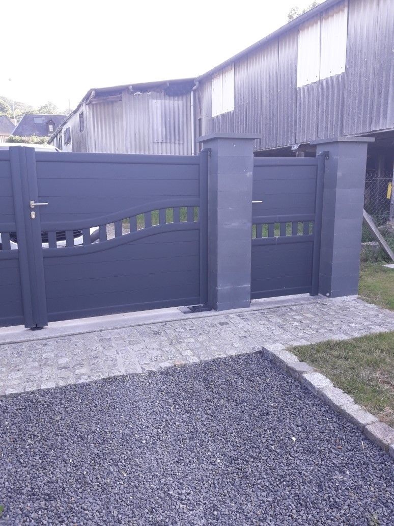 Portail en métal gris orné de découpes décoratives, sur un chemin de pierre, avec un bâtiment en arrière-plan.