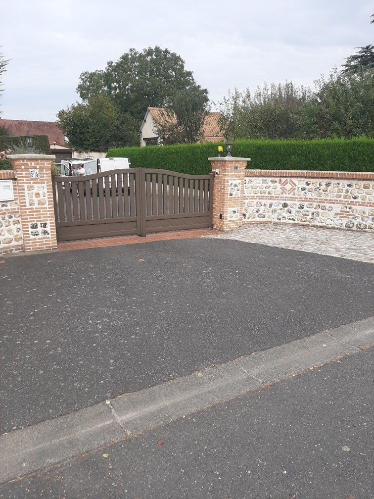 Portail en bois dans un mur de briques et de pierres, donnant sur une allée carrossable, avec une haie verte et des arbres en arrière-plan.