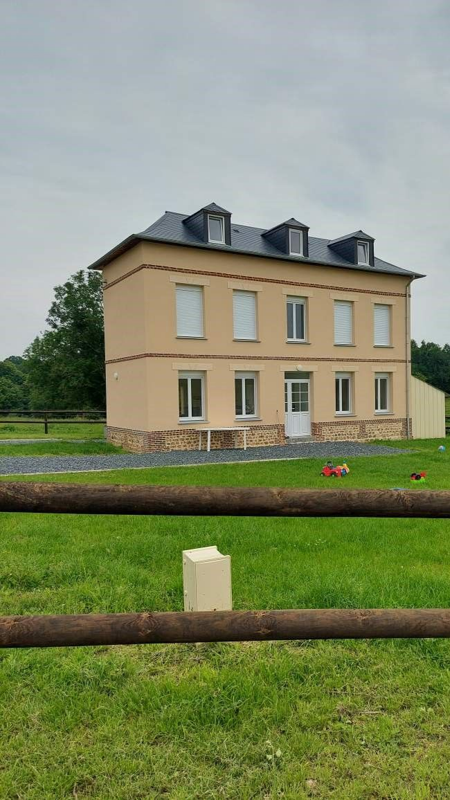 Maison beige à deux étages avec un toit gris et des lucarnes, située dans un champ herbeux avec une clôture en bois au premier plan.