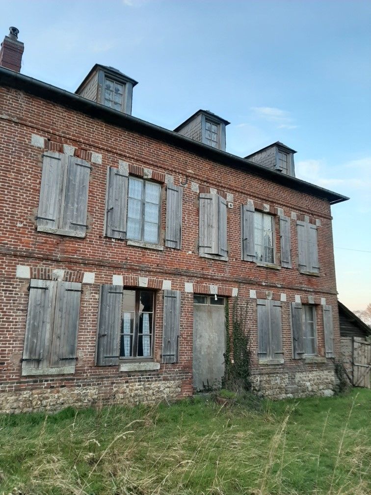 Maison en briques aux volets et lucarnes en bois patiné, située dans un champ herbeux sous un ciel nuageux.