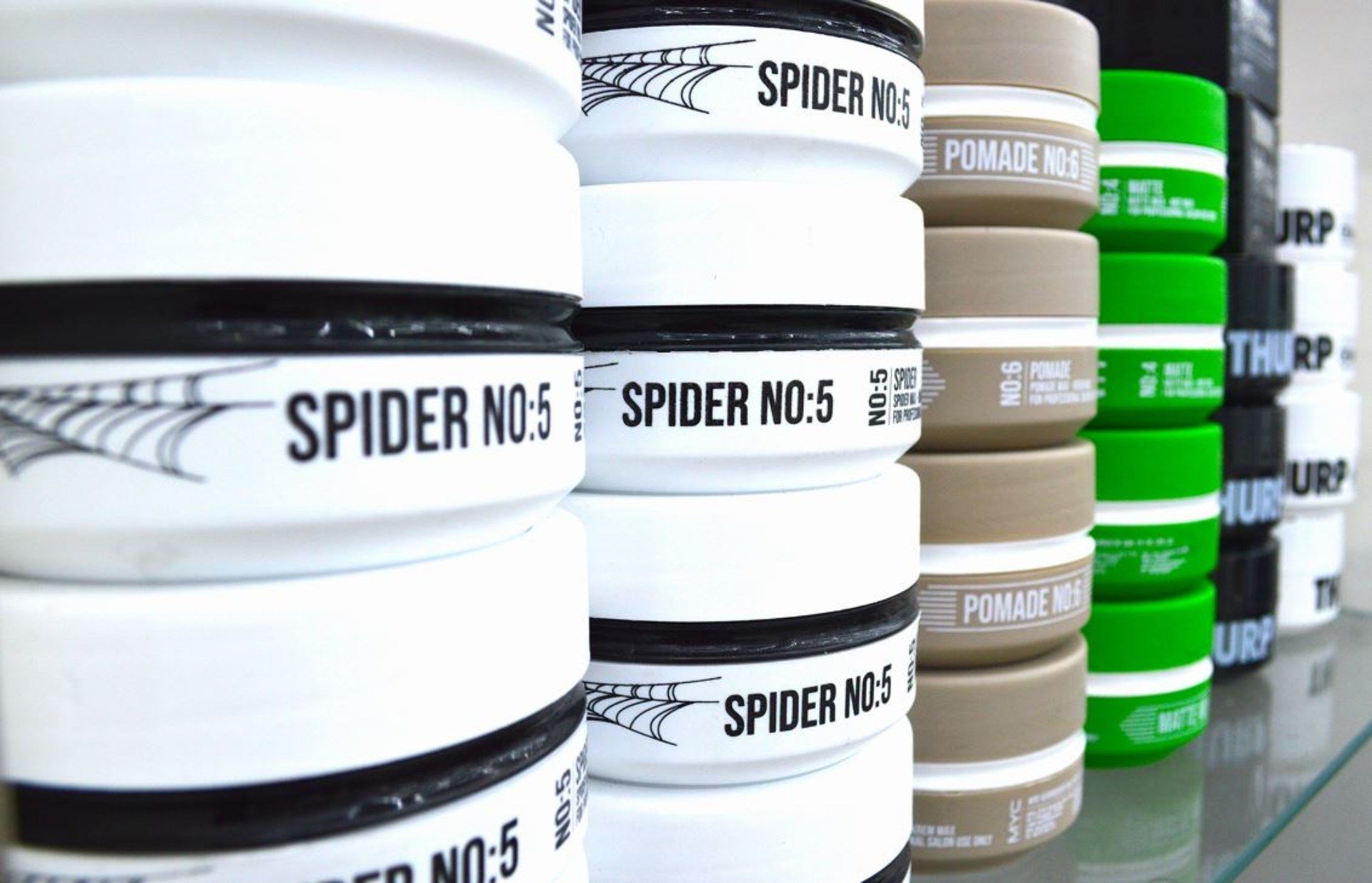 Auf einem Glasregal stapeln sich Behälter mit Spider No. 5, beiger Pomade und grünen Haarprodukten.