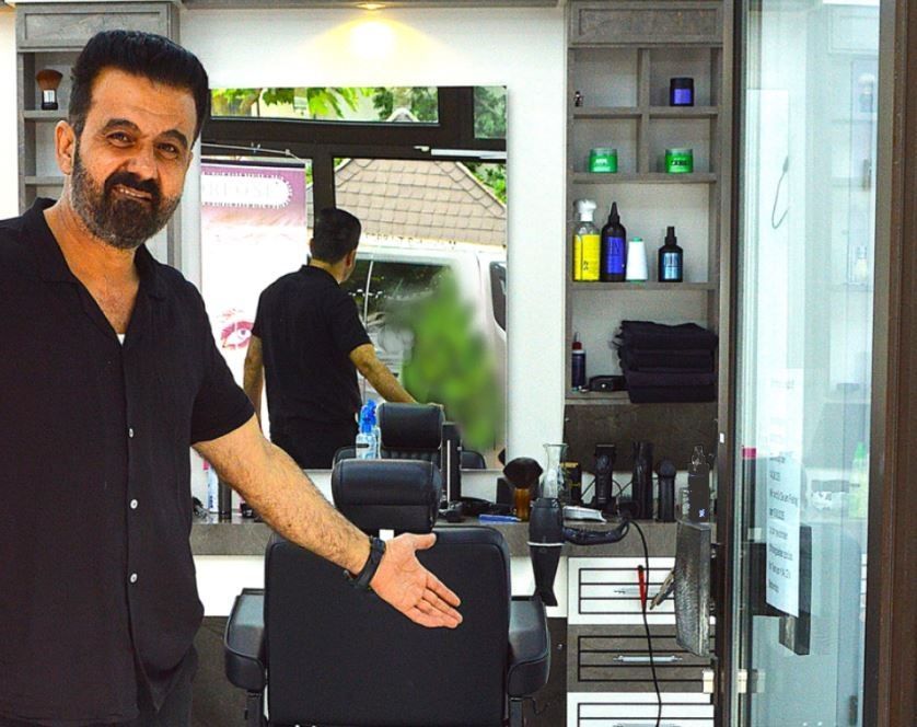 Ein Mann in schwarzem Hemd gestikuliert in einem modernen Friseursalon; hinter ihm befinden sich Spiegel, Stühle und Produktregale.
