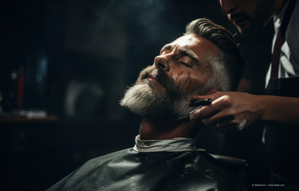 Bärtiger Mann lässt sich in einem düsteren Friseursalon die Haare schneiden, die Augen geschlossen, die Haarschneidemaschine in der Nähe seines Halses.