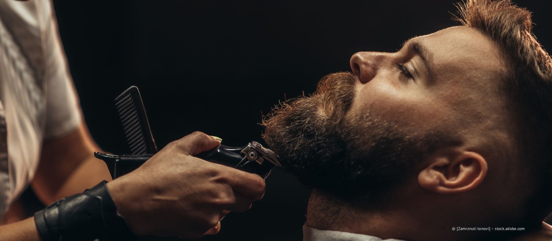 In einem dunklen, professionellen Ambiente stutzt ein Barbier den Bart eines Mannes mit einer elektrischen Haarschneidemaschine und einem Kamm.