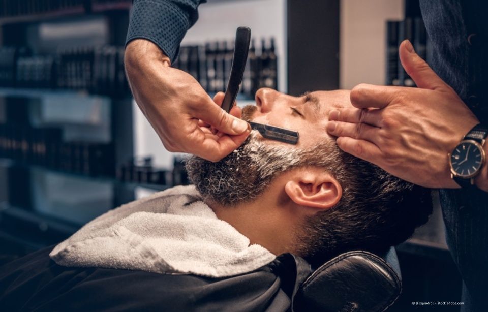 Ein Barbier rasiert einem Kunden in einem modernen Barbershop mit einem Rasiermesser den Bart.