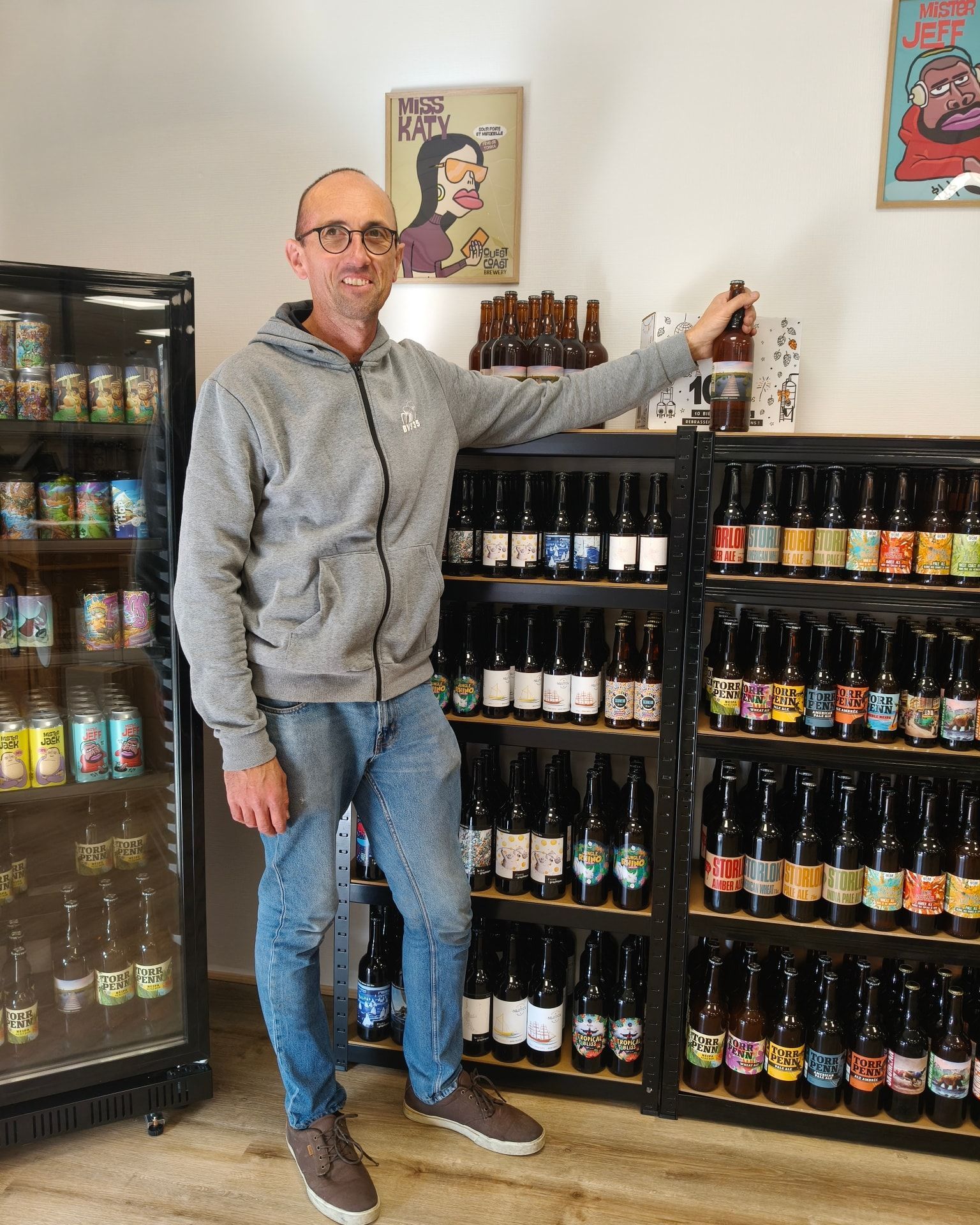 Un homme se tient dans une allée de magasin, près des étagères de boissons en bouteille, tenant une bouteille et souriant.