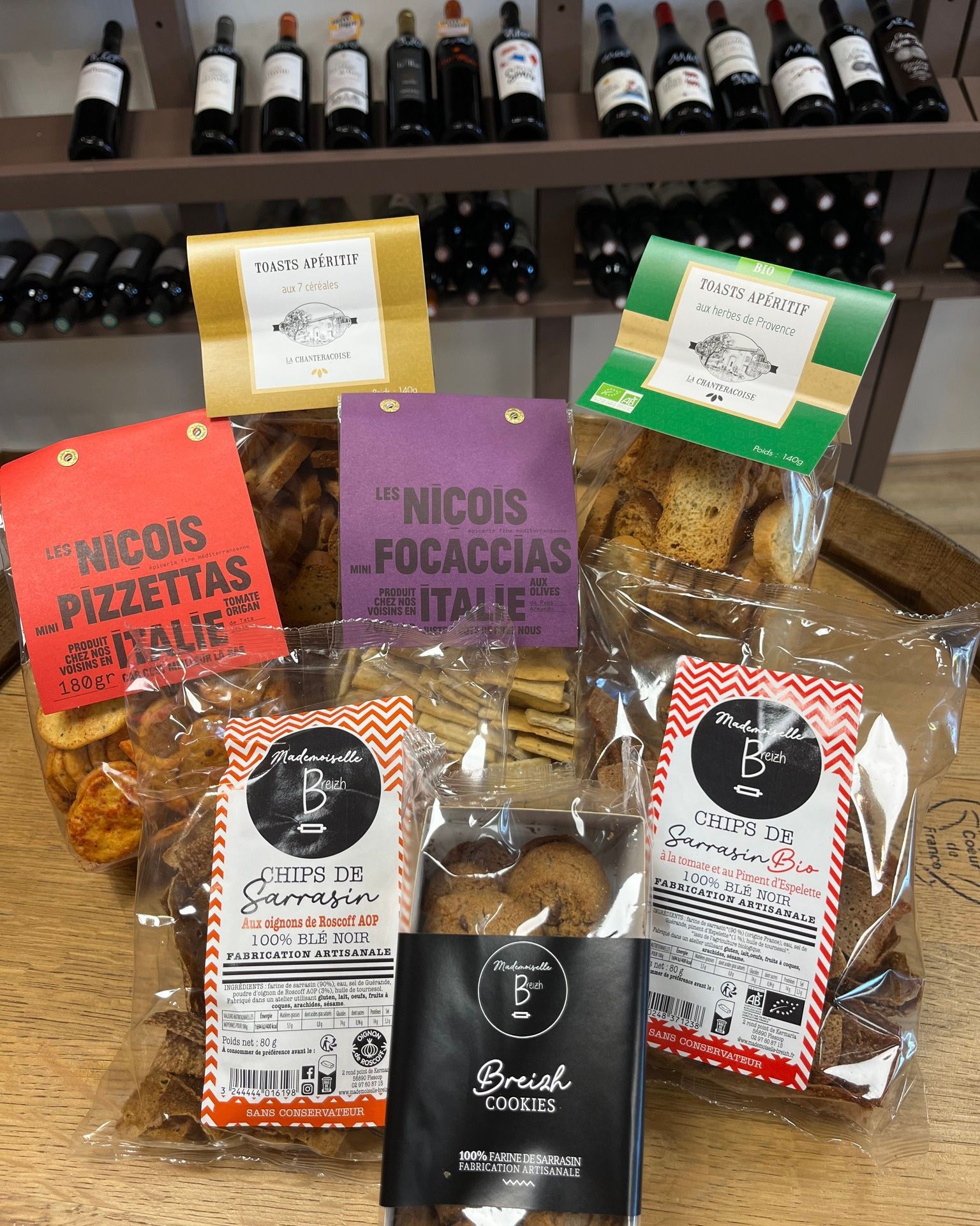 Des produits de boulangerie emballés assortis sont présentés sur une étagère, avec des bouteilles de vin en arrière-plan.