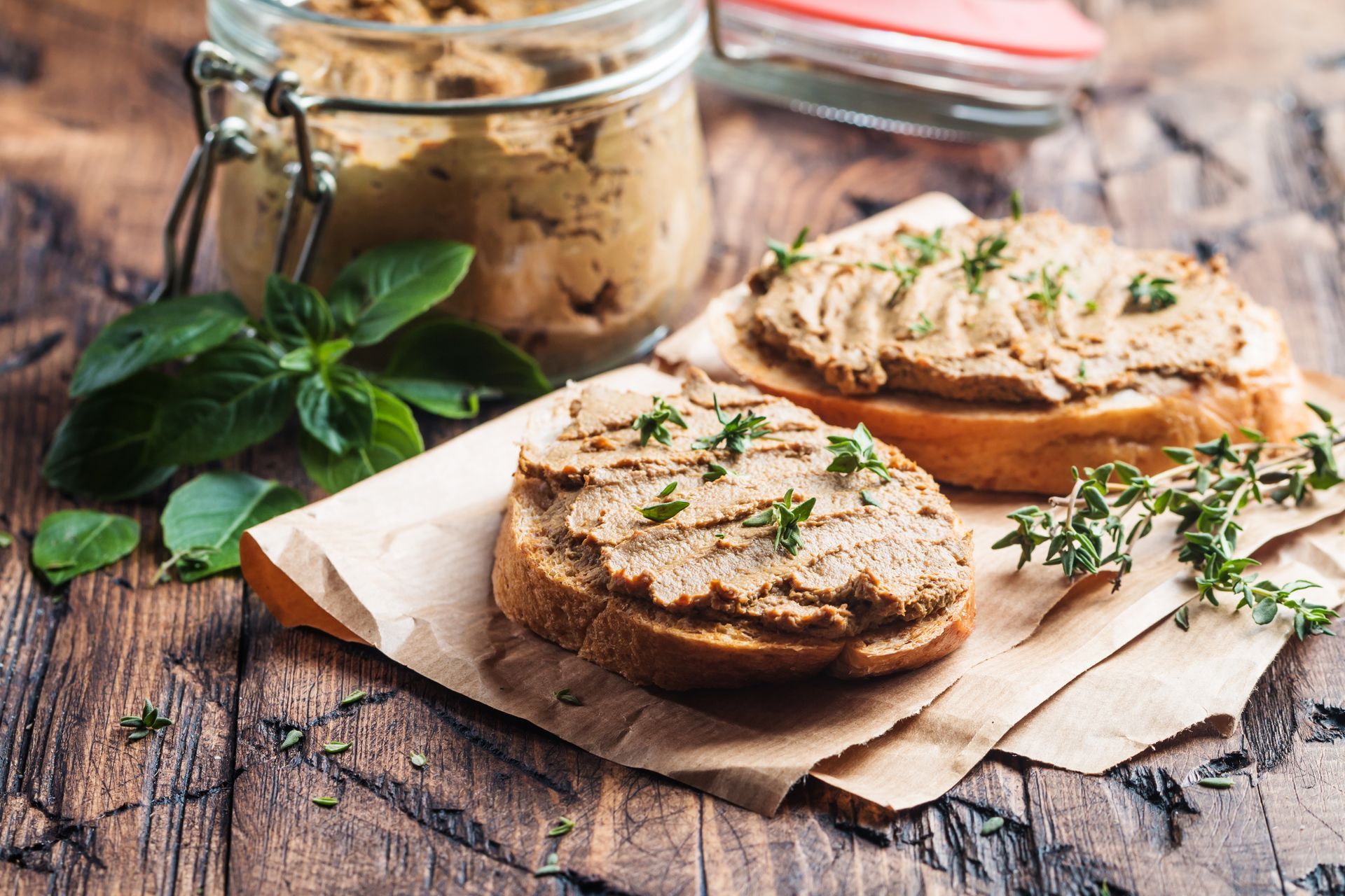 Des tranches de pain rustique tartinées d'herbes aromatiques, à côté de basilic frais et d'un bocal, sur une table en bois.