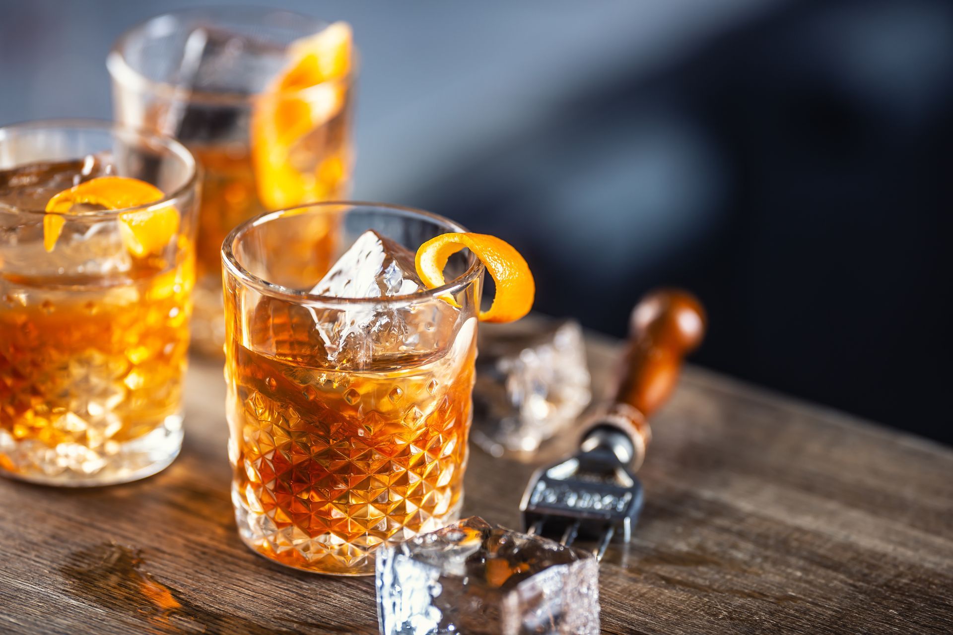 Deux cocktails au whisky, garnis d'orange, sont posés sur une table en bois, avec des glaçons, une bouteille et un tire-bouchon à proximité.