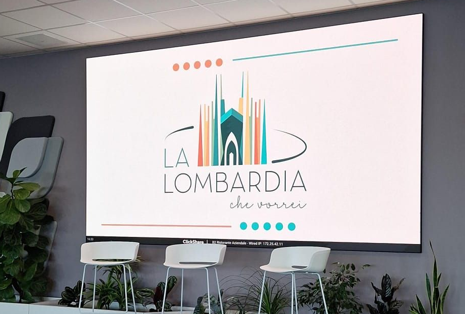 Palco moderno per eventi con il logo de La Lombardía su un grande schermo e sedie bianche di fronte.