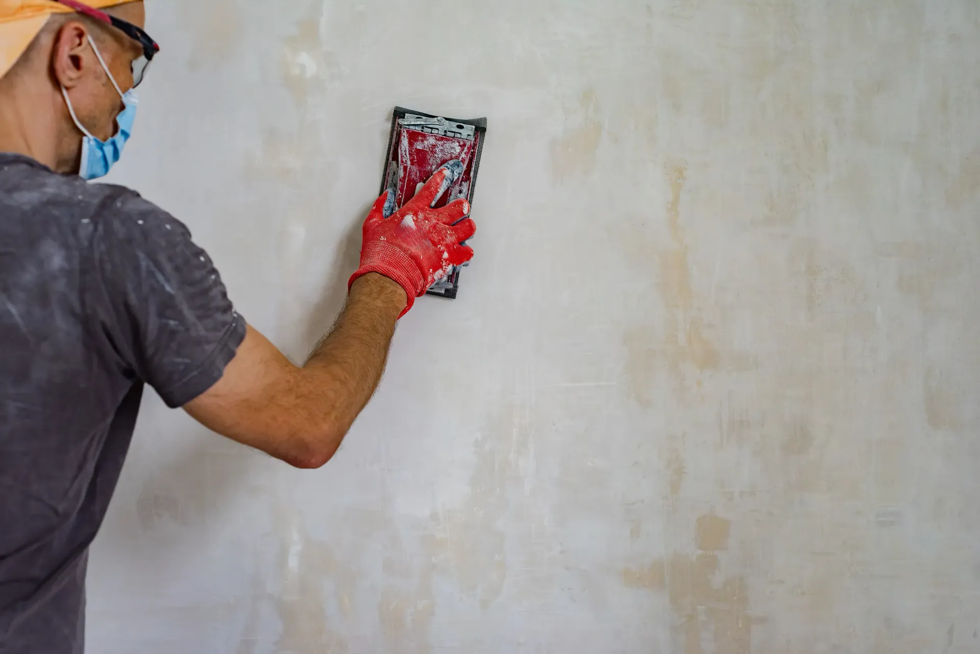 Un trabajador con mascarilla y guantes rojos utiliza un taco de lija para alisar una pared enlucida.
